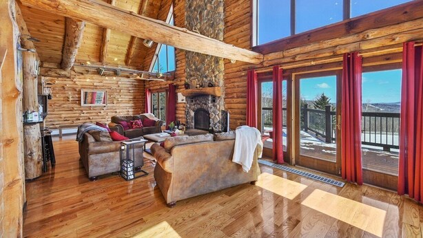 A log cabin interior with a fireplace, wooden floors, and large windows offering a view of the outdoors. - Berkshire Mountain Top Chalet (North Adams)