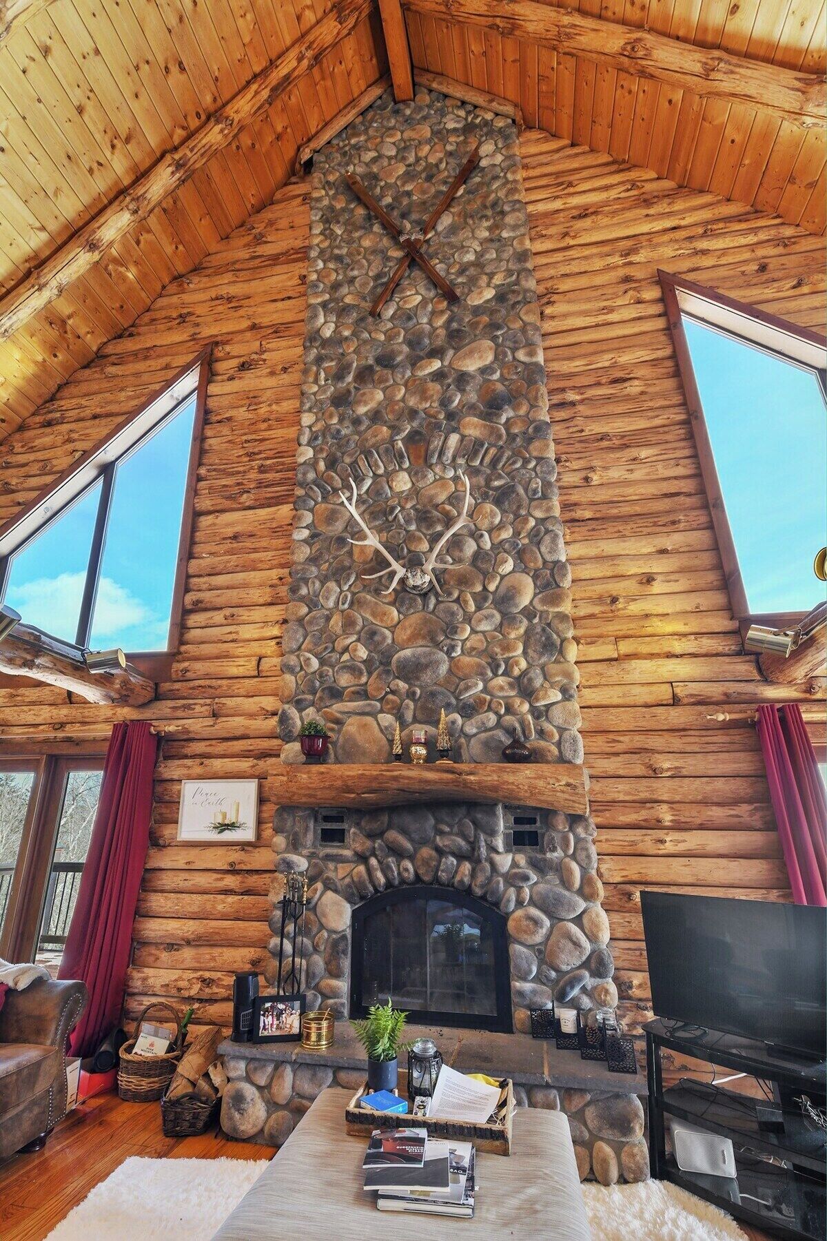 A log cabin interior with a stone fireplace, wooden walls, and a large window.