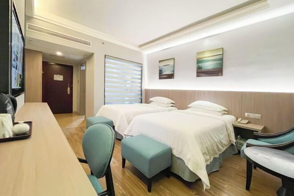 Habitación de hotel con dos camas, un escritorio y una silla. Cuenta con una ventana con persianas, un cuadro en la pared y una puerta que da a otra habitación.