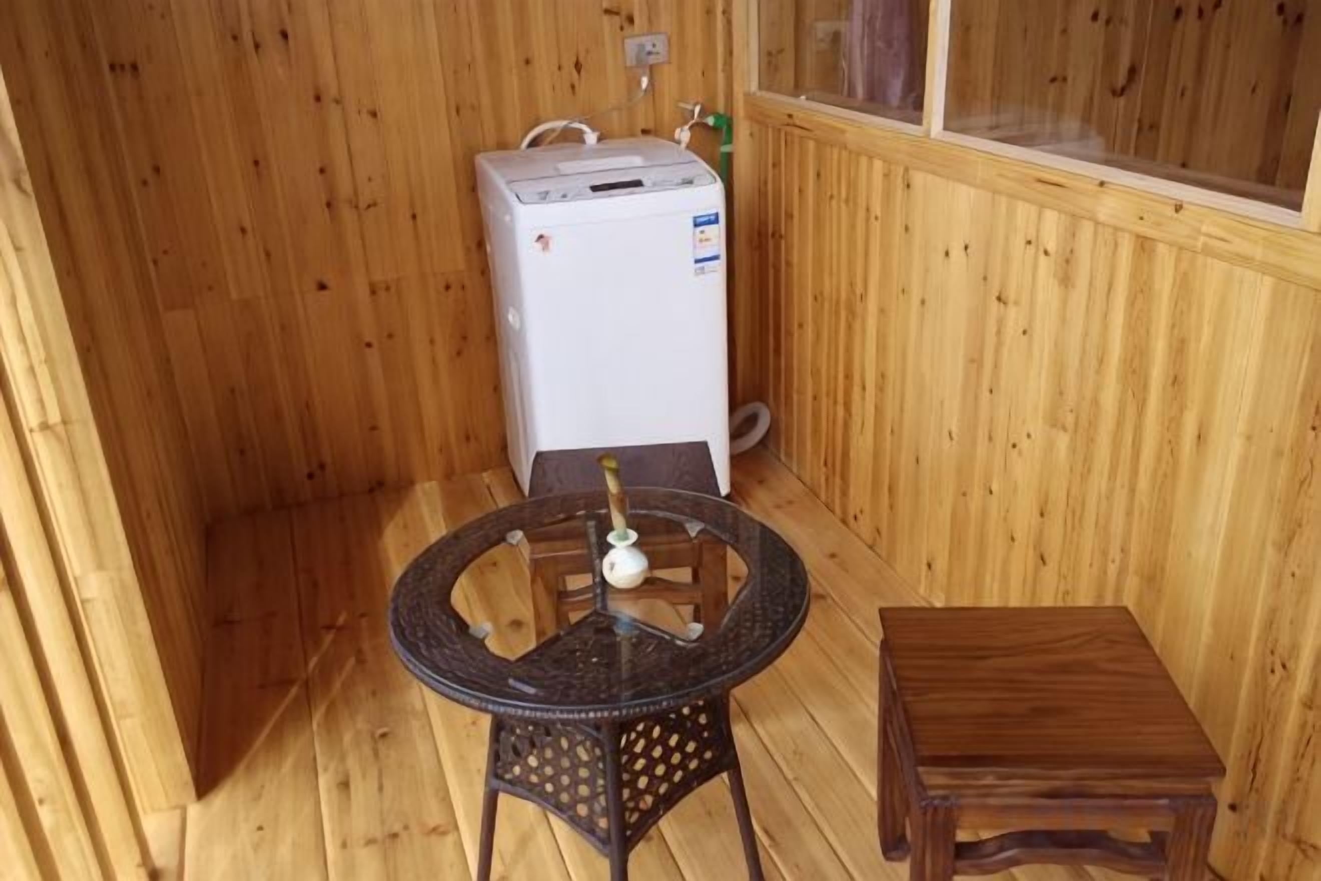 Quarto pequeno com paredes de madeira, um aquecedor branco, uma mesa redonda com uma garrafa e um banquinho de madeira.
