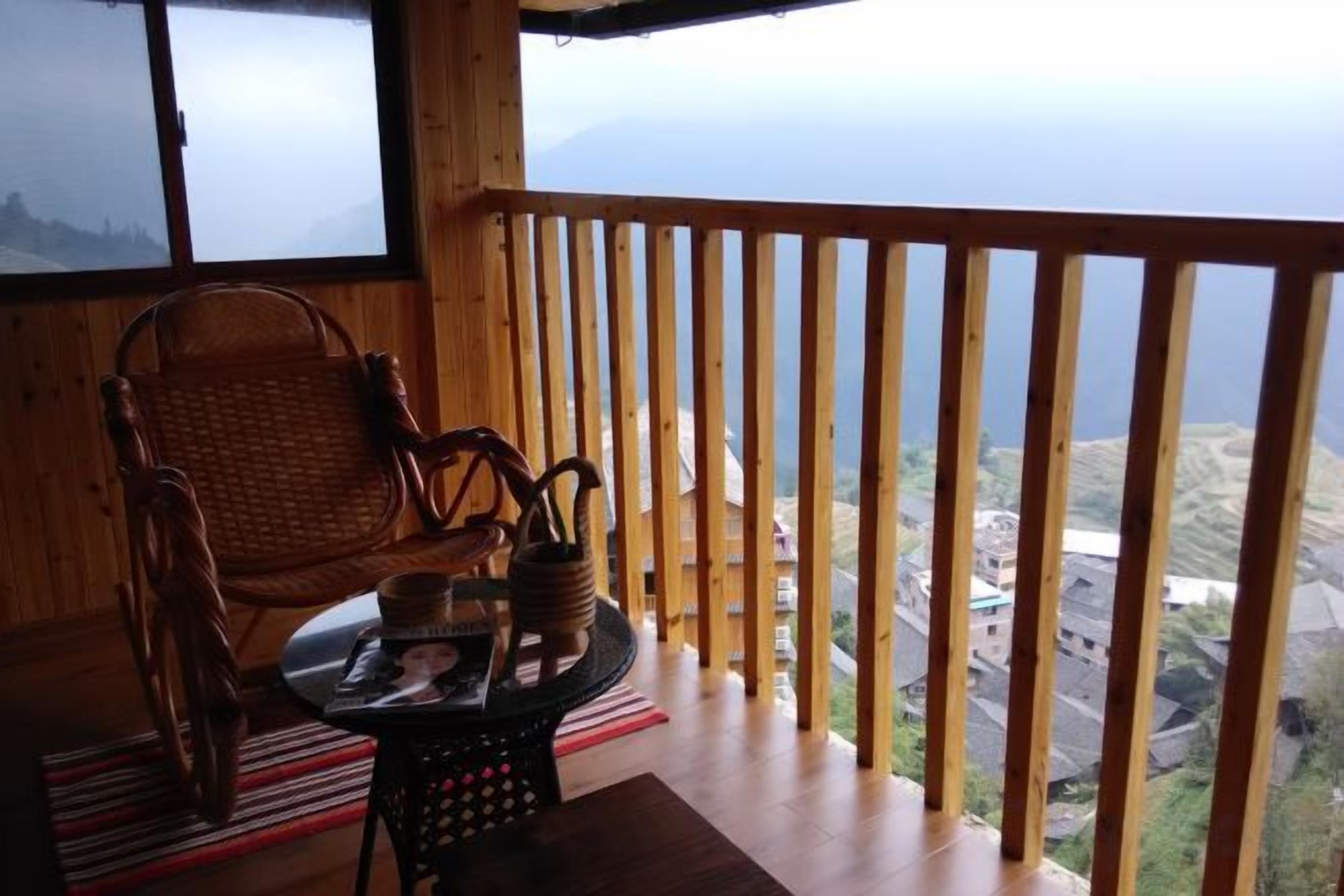A wooden balcony with a wicker chair and a small table with a tray, overlooking a mountainous landscape.