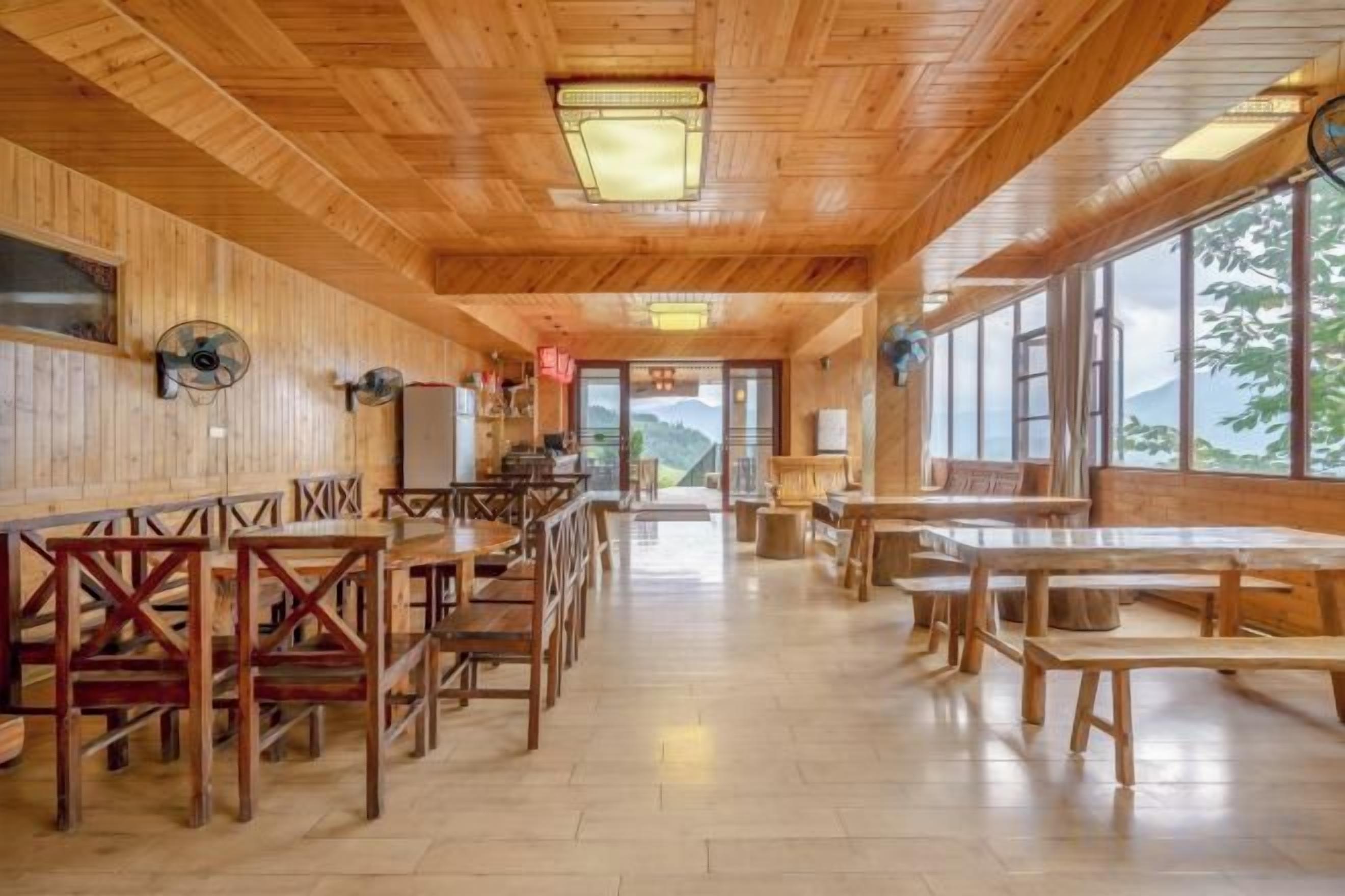 A wooden interior with tables and chairs arranged for dining, large windows offering a view, and a ceiling fan.
