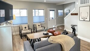 A modern living room with a sofa, armchairs, a coffee table, and a TV. There are windows with blinds, a staircase, and a door. - Pristine Home Near Sun Valley (Hailey)