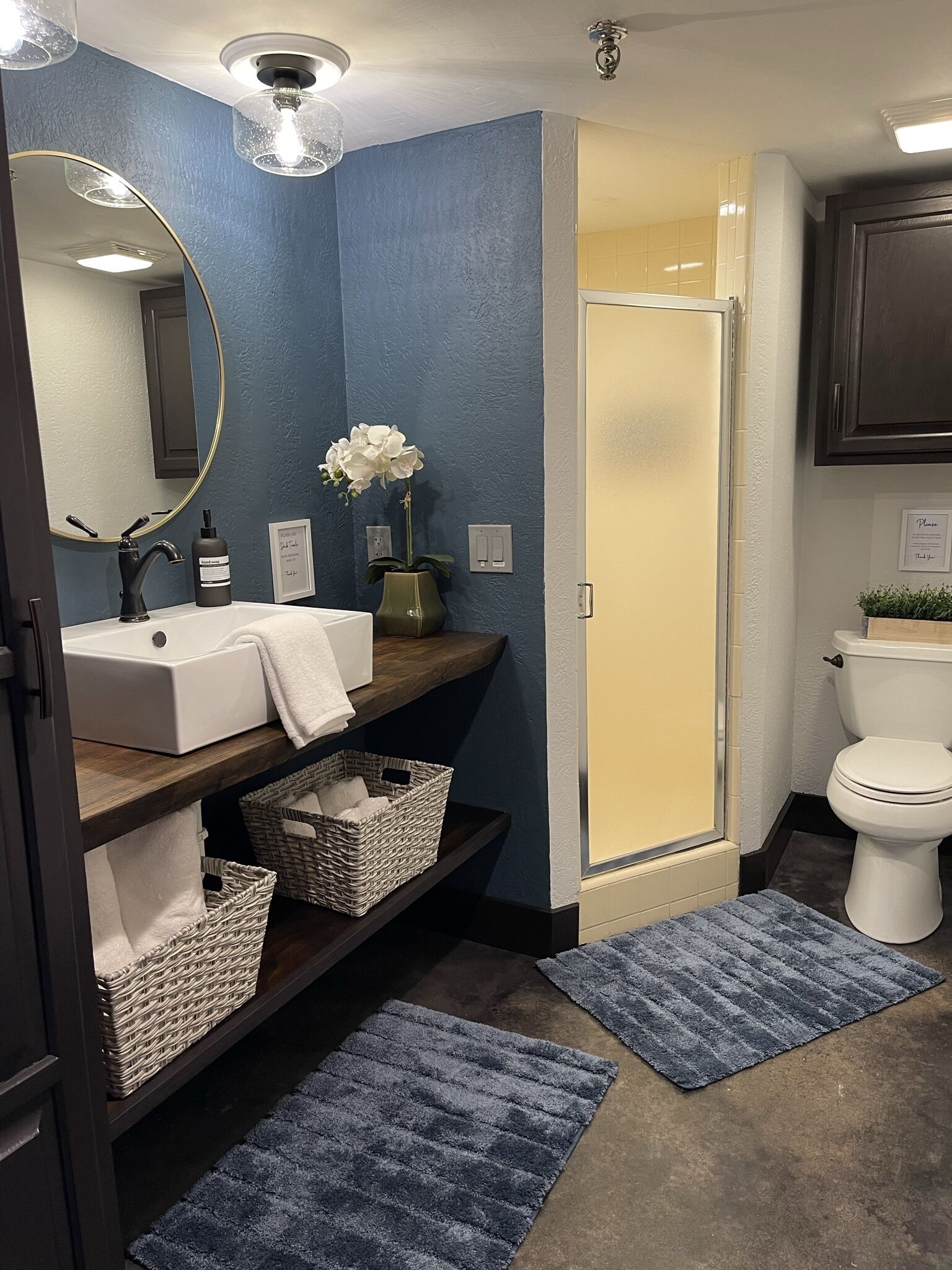Une salle de bain avec un lavabo blanc, un miroir, une douche et des toilettes. Il y a deux paniers sous le lavabo et un vase avec des fleurs sur le comptoir.
