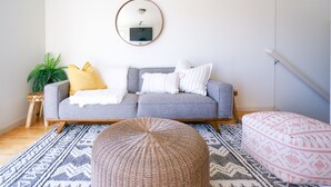 Ein Wohnzimmer mit grauem Sofa, einem runden Hocker, einem gemusterten Teppich und einer Topfpflanze.