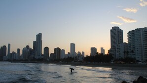 A coastal cityscape with high-rise buildings, a beach, and a person carrying a surfboard.