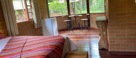 A cozy room with a bed, a small table, and chairs, a refrigerator, and a view of greenery outside.