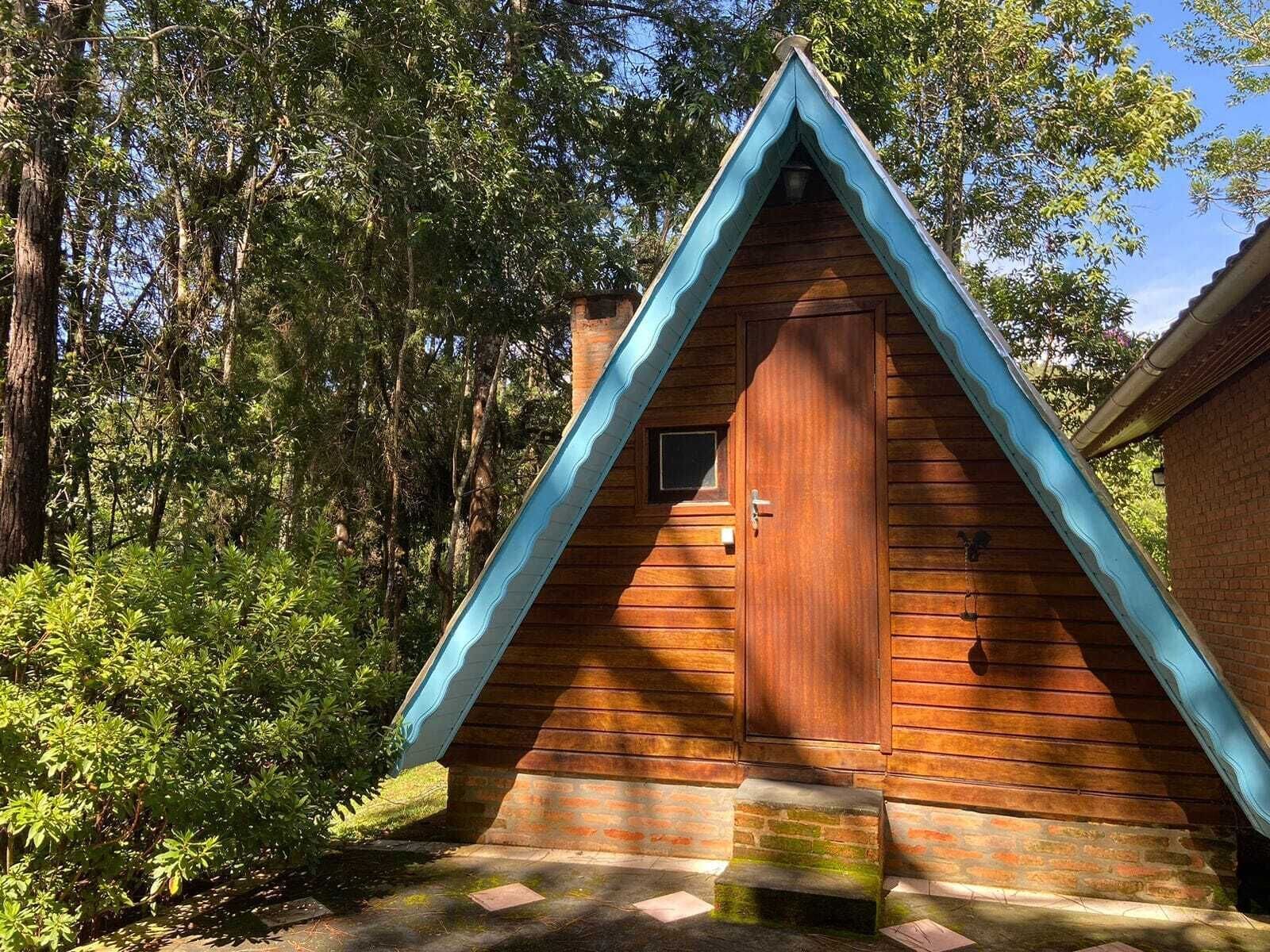 A wooden cabin with a triangular roof and a door, surrounded by trees and greenery.