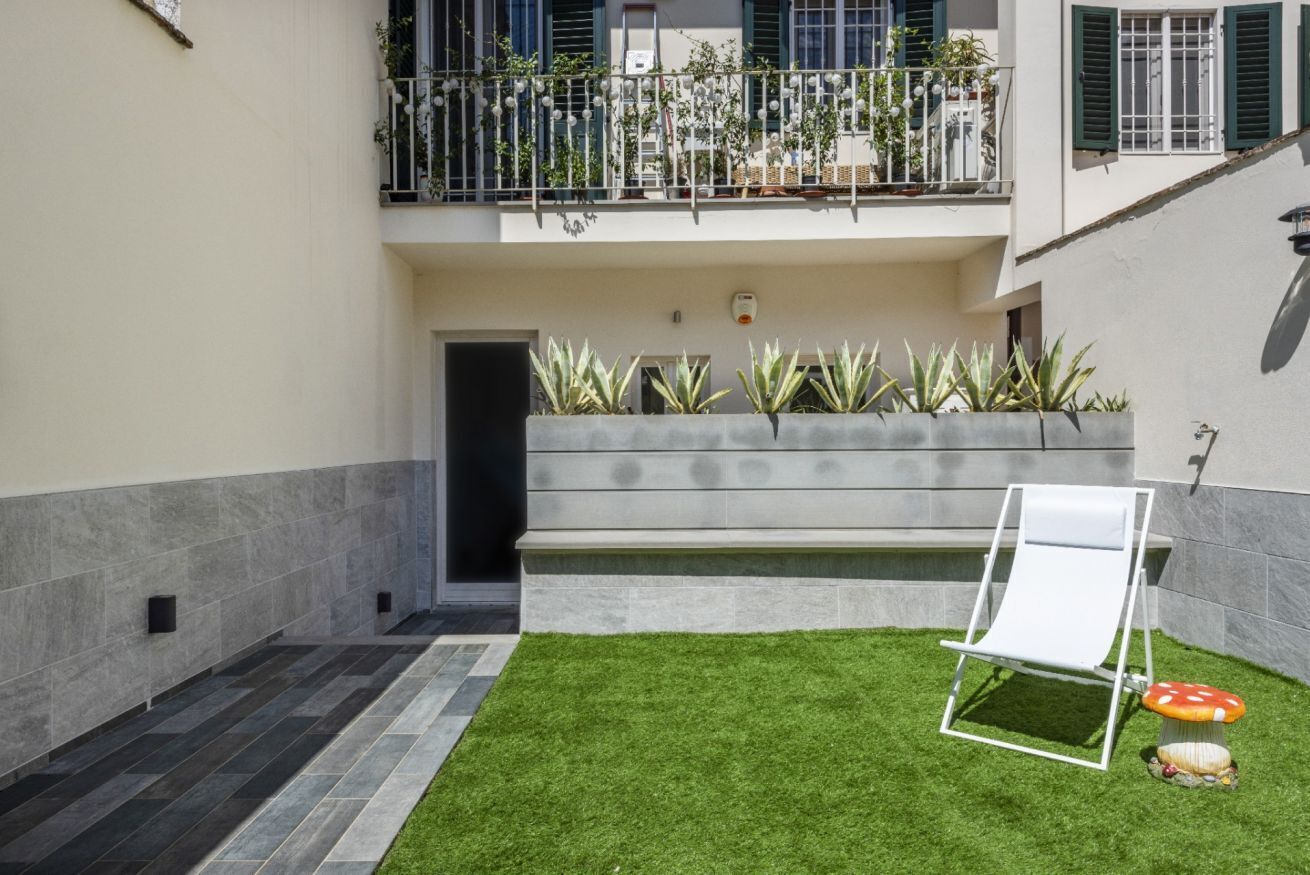 A courtyard with a white lounge chair, a small garden with green grass and plants, and a balcony with a railing and potted plants.
