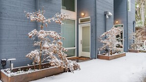 En modern byggnad med en blå fasad, en entré med glasdörr och snötäckta växter i förgrunden.