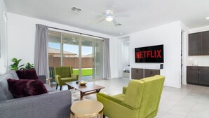 Una sala de estar moderna con un sofá gris, un sillón verde y una mesa de centro. Un televisor de pantalla plana grande está montado en la pared mostrando el logotipo de Netflix.