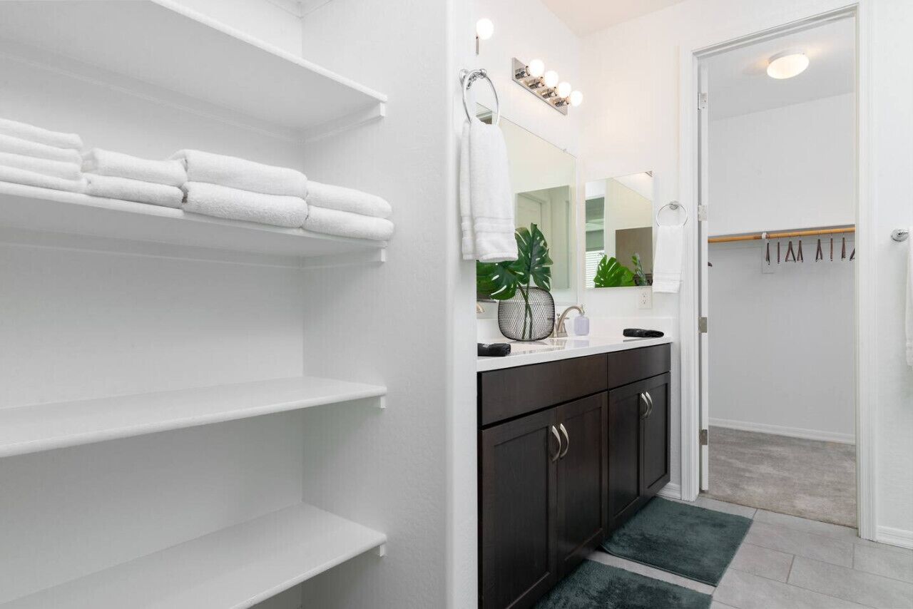 Une salle de bain avec des étagères blanches, un meuble sombre, un miroir avec éclairage intégré et une plante verte.