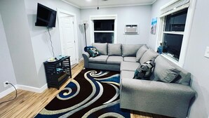 A modern living room with a grey sofa, a black and blue rug, a ceiling fan, and a flat-screen TV mounted on the wall. - The Bungalow on Betty (Clearwater)