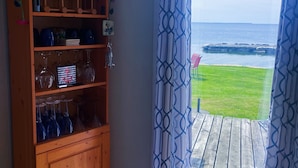 Een houten kast met wijnglazen en planten in potten, uitzicht op de oceaan door gordijnen met patroon, en een houten terras.