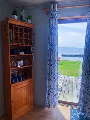 A wooden cabinet with wine glasses and potted plants, a view of the ocean through patterned curtains, and a wooden deck.