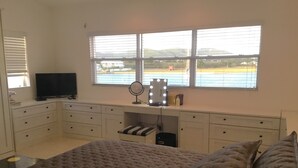 Un dormitorio con cama, escritorio con espejo e iluminación, televisor y ventana con vistas a un campo y colinas.