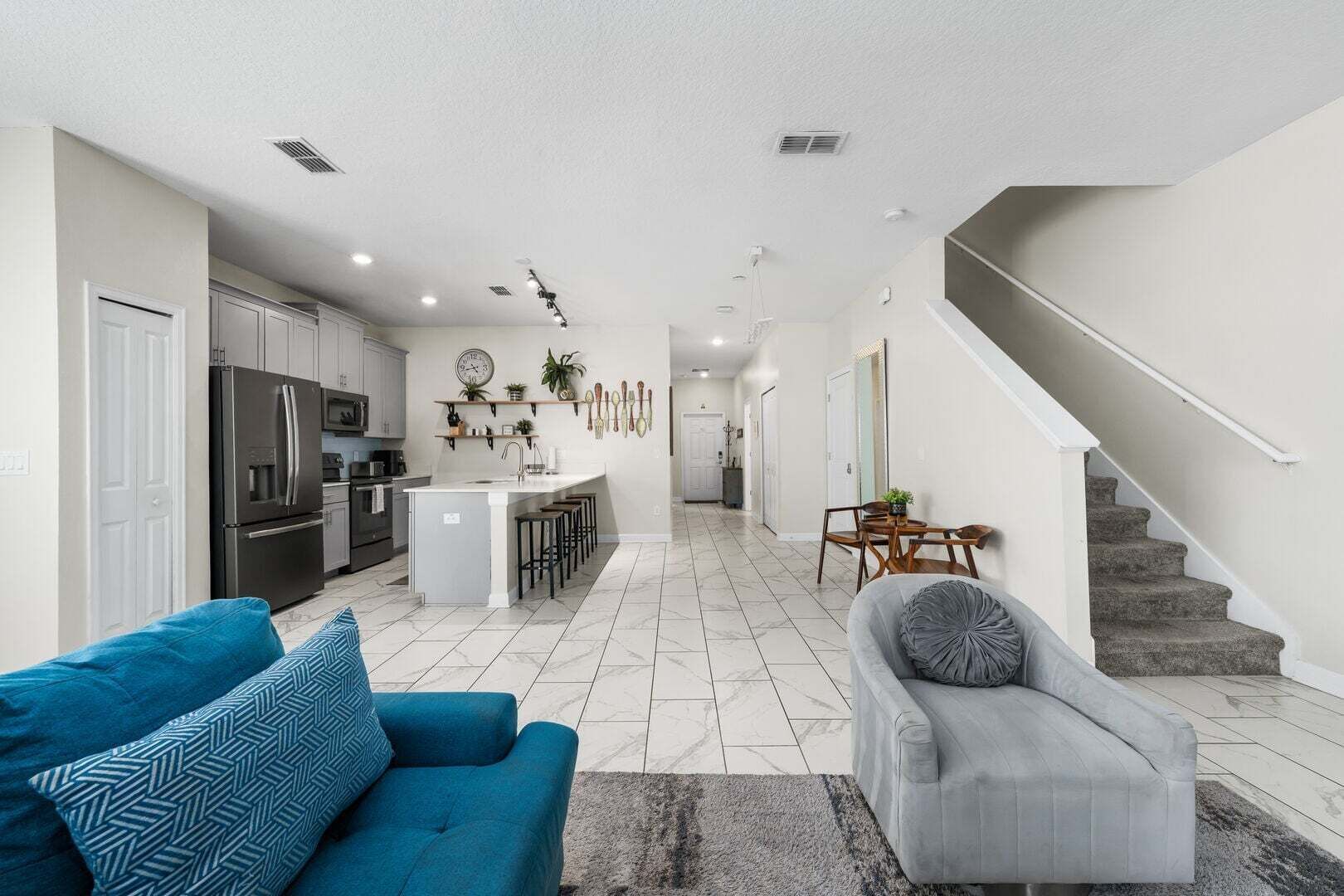 A modern kitchen with a central island, a dining area with a table and chairs, a living room with a sofa and a chair, and a staircase.