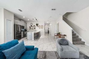 A modern kitchen with a central island, a dining area with a table and chairs, a living room with a sofa and a rug, and a staircase leading to another level.