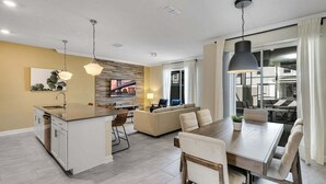 A modern kitchen with a central island, a dining area with a wooden table and chairs, and a living area with a sofa and TV. - Stylish Family Home | Private Pool | 111 (Kissimmee)