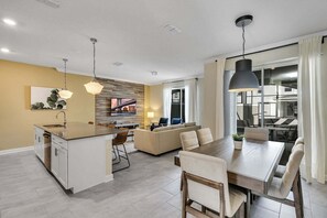A modern kitchen with a central island, a dining area with a large table, and a living area with a sofa and TV.