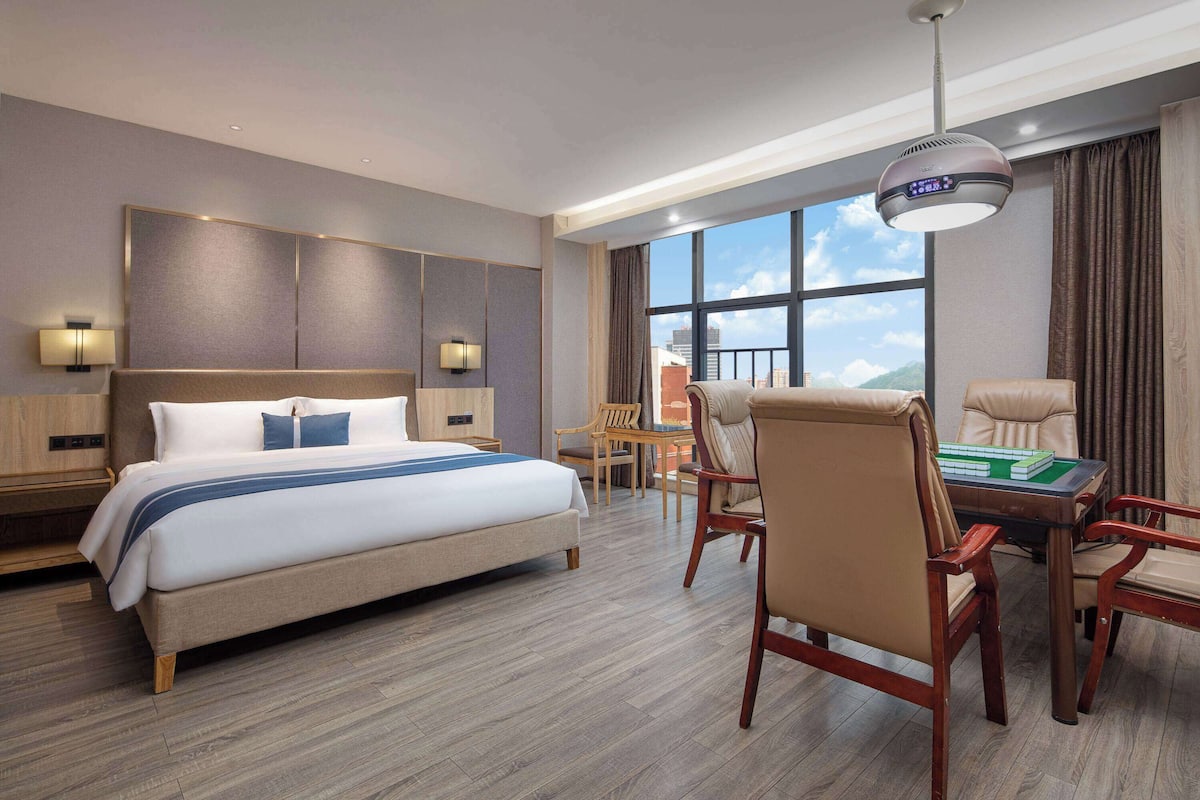 Quarto de hotel com uma cama grande, uma escrivaninha com cadeiras, vista para a cidade e uma luminária no teto.