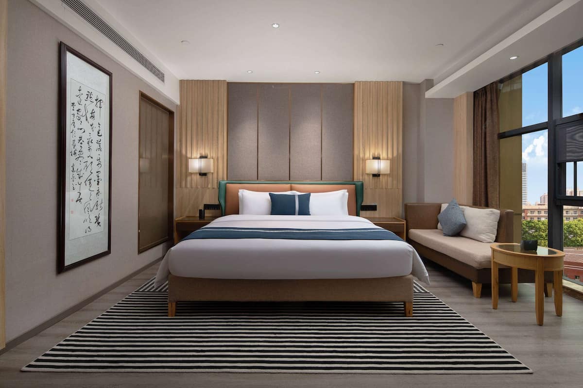 Quarto de hotel moderno com uma cama grande, um sofá, uma mesinha e uma janela ampla com vista para a cidade.