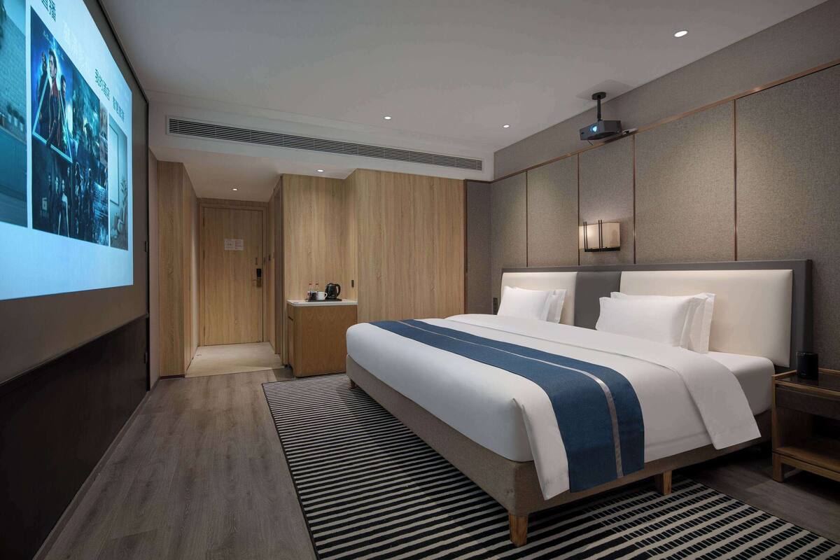 Quarto de hotel moderno com uma cama grande, mesa de cabeceira, guarda-roupa de madeira e um projetor.