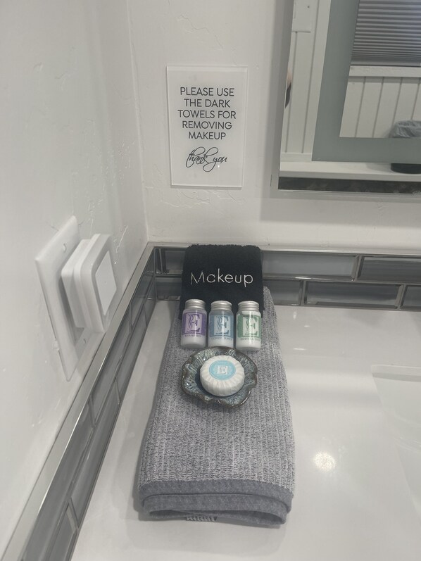 A bathroom with a sign instructing to use dark towels for makeup removal. There are three bottles of shampoo, body lotion, and conditioner on a towel labeled 'Makeup.'