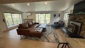 Amplia sala de estar con sofá de cuero, chimenea de piedra y ventanas grandes con vistas al exterior.