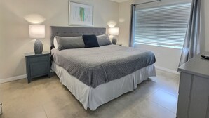 Un dormitorio con cama, dos lámparas de noche, ventilador de techo, ventana con persianas y un cuadro en la pared.