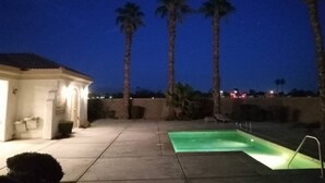 A residential area with a swimming pool, palm trees, and a house with exterior lighting.