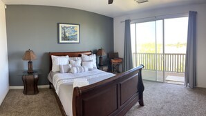 A bedroom with a large bed, two bedside tables, a chair, a painting on the wall, and a balcony with a view.