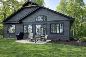 Une maison moderne à deux étages, avec une façade de couleur gris foncé, de grandes fenêtres et un patio couvert aménagé avec du mobilier d’extérieur.