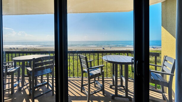 A balcony with a view of the beach, featuring outdoor furniture and a glass door. - Oceanfront Luxury Condo - 3BD with Gorgeous Views - Oceanfront Pool (North Myrtle Beach)