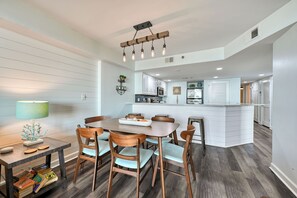 A dining area with a wooden table and chairs, a kitchen in the background, and a modern pendant light fixture.