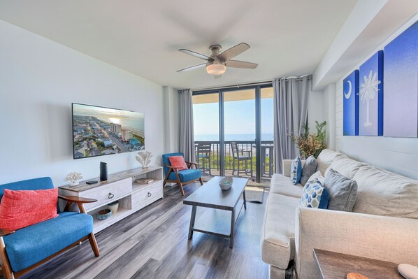 A modern living room with a large TV, blue and red chairs, a grey sofa, and a balcony with a view of the city.
