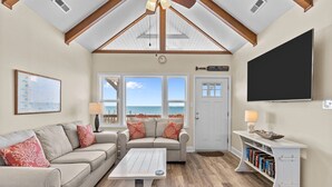 Una sala de estar acogedora con un sofá, una mesa de centro y una estantería. Amplias ventanas ofrecen vistas a la playa.