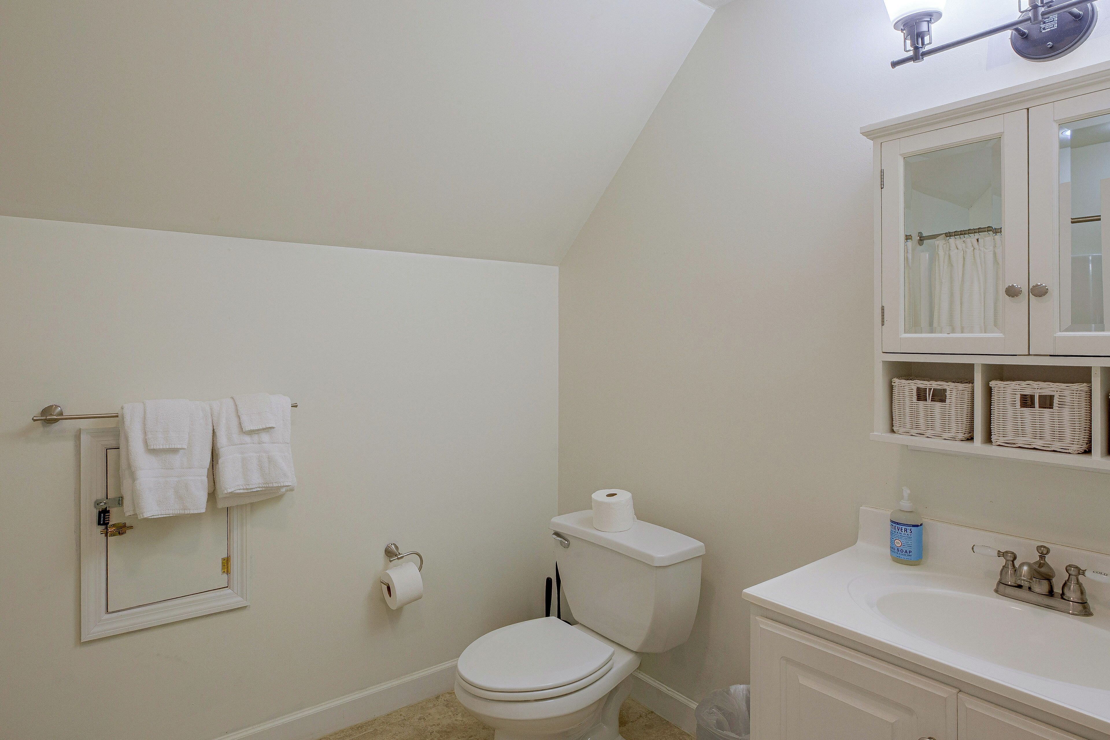 A bathroom with a toilet, a towel rack with towels, a toilet paper holder, a sink with a soap dispenser, and a mirror cabinet.