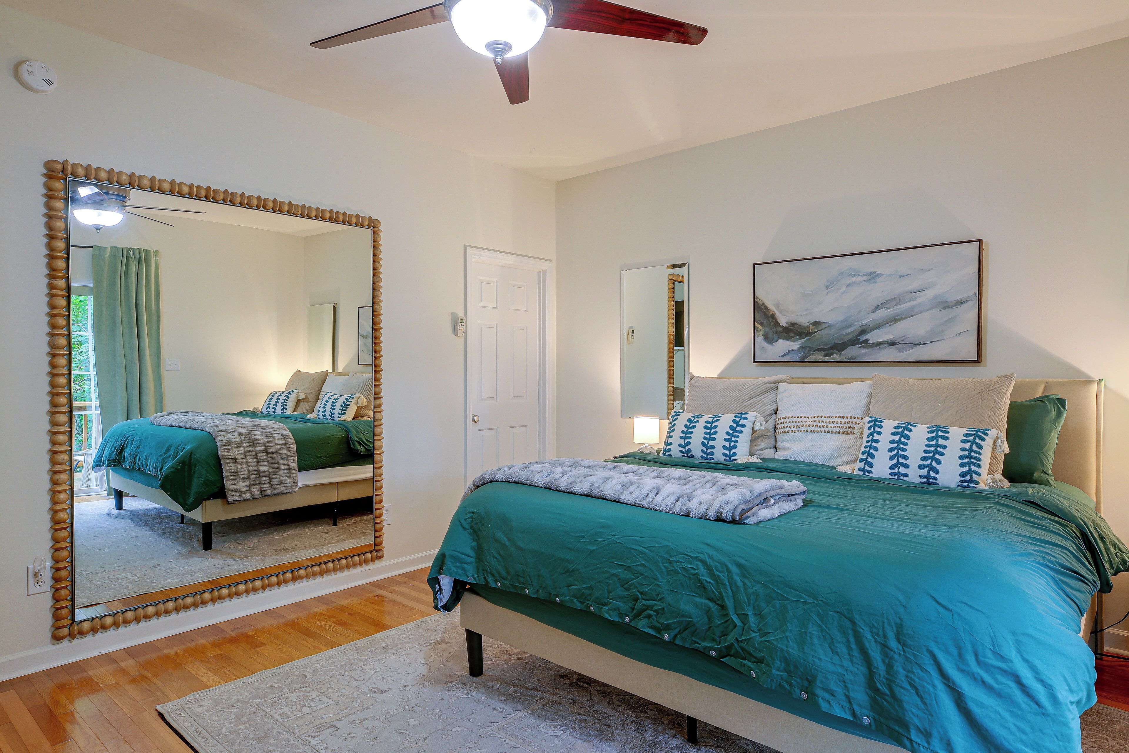 A bedroom with a large mirror, a bed with green bedding, a ceiling fan, and a painting on the wall.