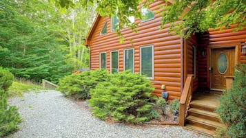 A log cabin with a gravel driveway, surrounded by dense greenery and trees.