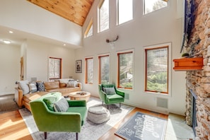 Een ruime woonkamer met een hoge plafond, grote ramen, een stenen openhaard en een gezellige zithoek met een groene fauteuil en een bank.