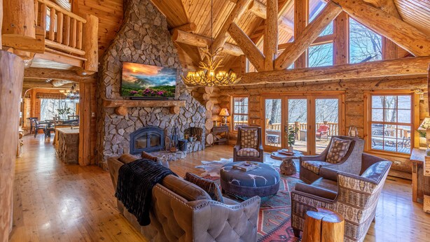A spacious log cabin interior with a stone fireplace, wooden beams, and large windows.