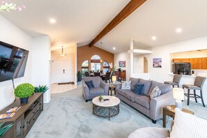 A modern living room with a flat-screen TV, sofas, a coffee table, and a kitchen area with wooden cabinets and a refrigerator.