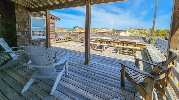 A wooden deck with Adirondack chairs and picnic tables, overlooking a beach and dunes.