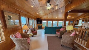 A cozy cabin interior with wicker furniture, a fireplace, and large windows offering views of the outdoors.