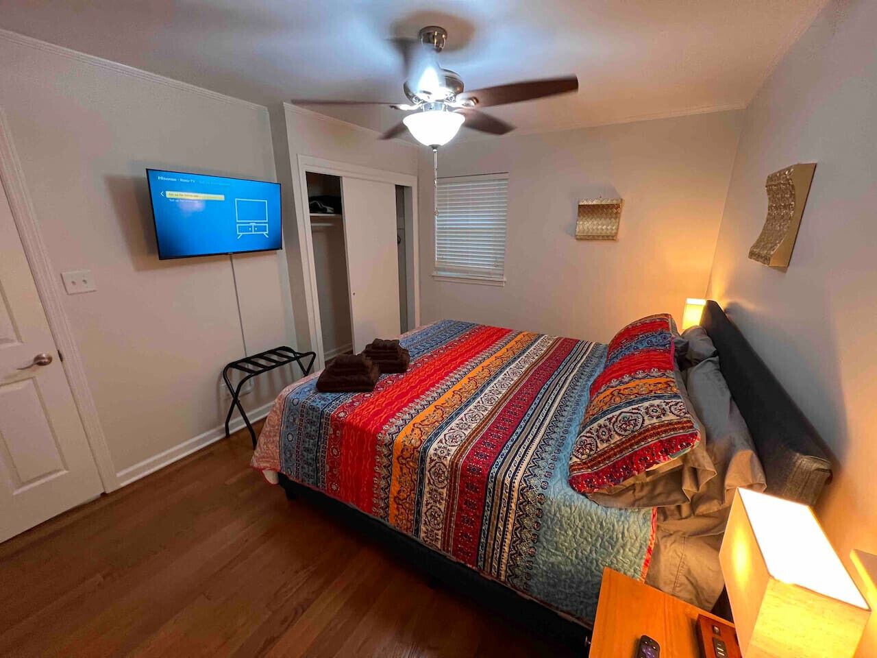 A bedroom with a bed, a ceiling fan, a television, and a nightstand with a lamp.