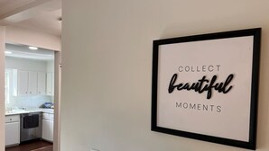 Une citation encadrée est accrochée au mur d’une cuisine moderne, avec des meubles blancs et un plancher en bois foncé.