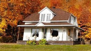 A two-story house with a front porch, surrounded by autumn foliage. - Les Chalets Tourisma - La Campagnarde (Saint-Leonard-de-Portneuf)