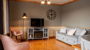 A living room with a grey sofa, a pink armchair, a TV on a white cabinet, and a floor lamp. - Les Chalets Tourisma - La Campagnarde (Saint-Leonard-de-Portneuf)