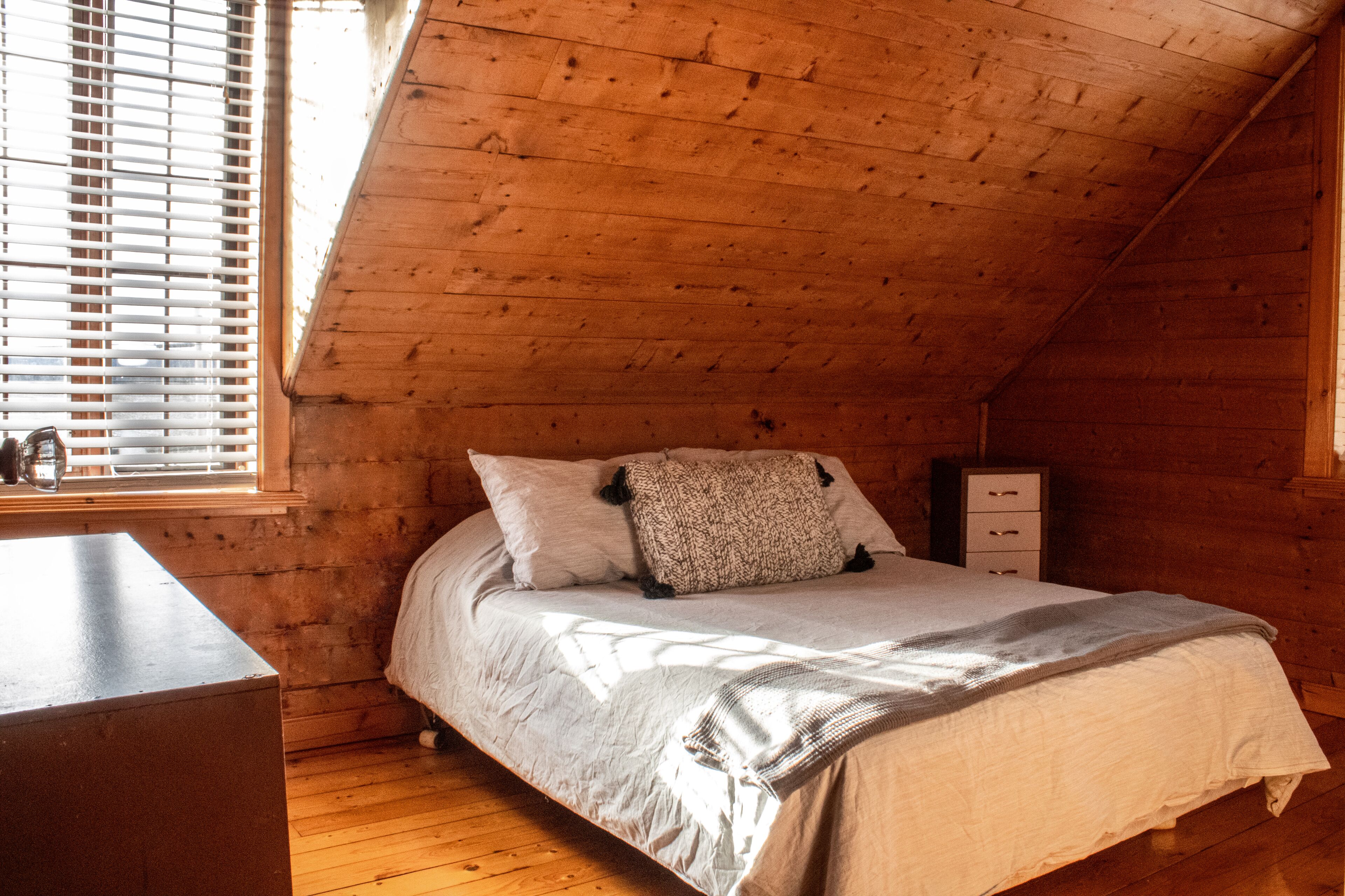 Ein Schlafzimmer mit schrägen Wänden, Holzwänden, einem Bett mit weißer Bettwäsche, einem Fenster mit Jalousien und einem Nachttisch.
